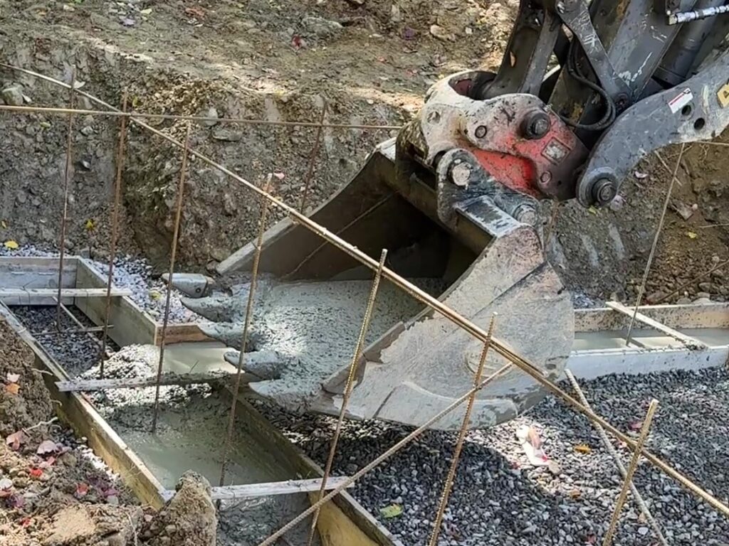 Excavator bucket tipping concrete into the rebar-lined footing forms for the Juniper Ridge cabin near Jefferson, NY, September 2026