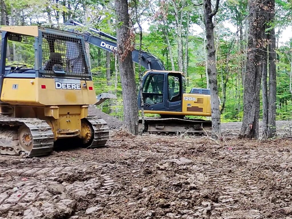 Yellow bulldozer and excavator on the graded, track-marked building site among tall hardwoods at Juniper Ridge on Blenheim Hill, NY, September 2026