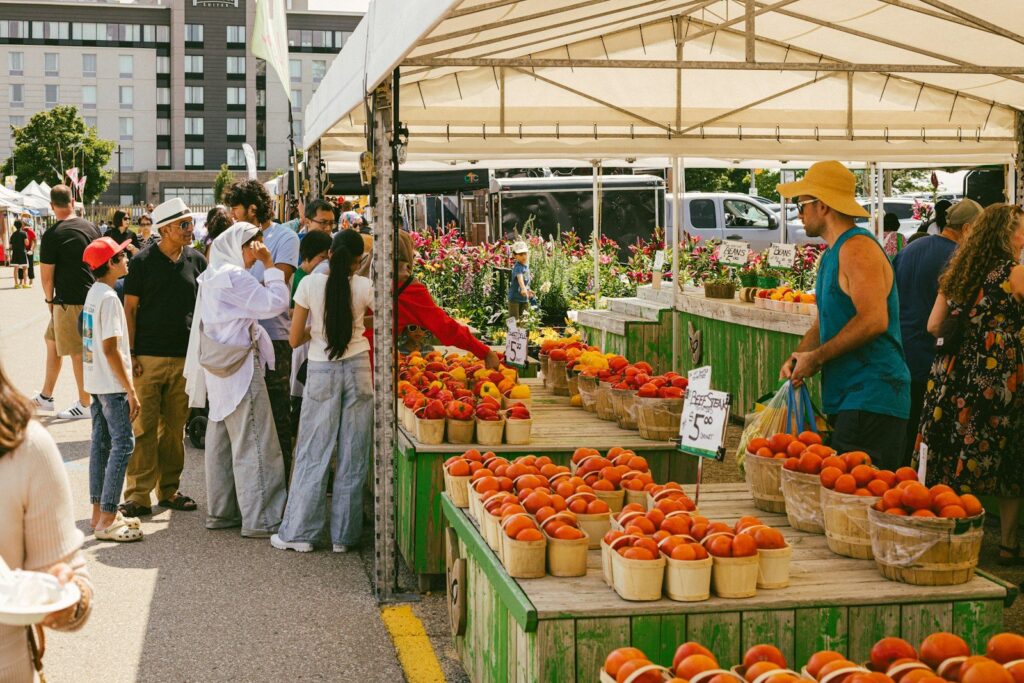 Shoppers at an outdoor farmers market stall piled with baskets of peaches and tomatoes